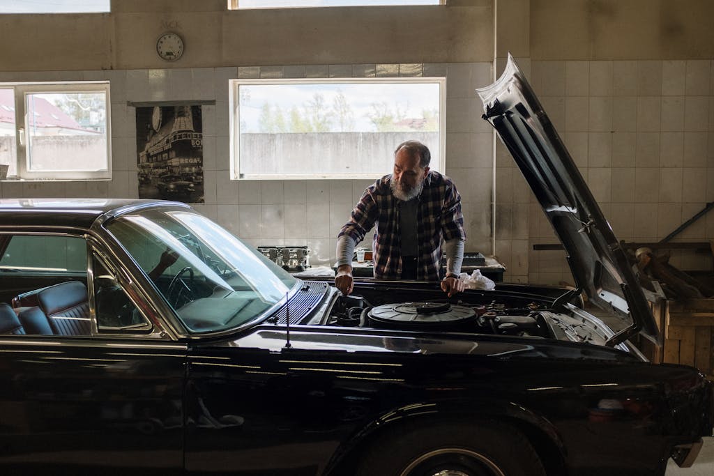 Imagen actual: A bearded mechanic examines a classic car engine in a well-lit auto repair shop.