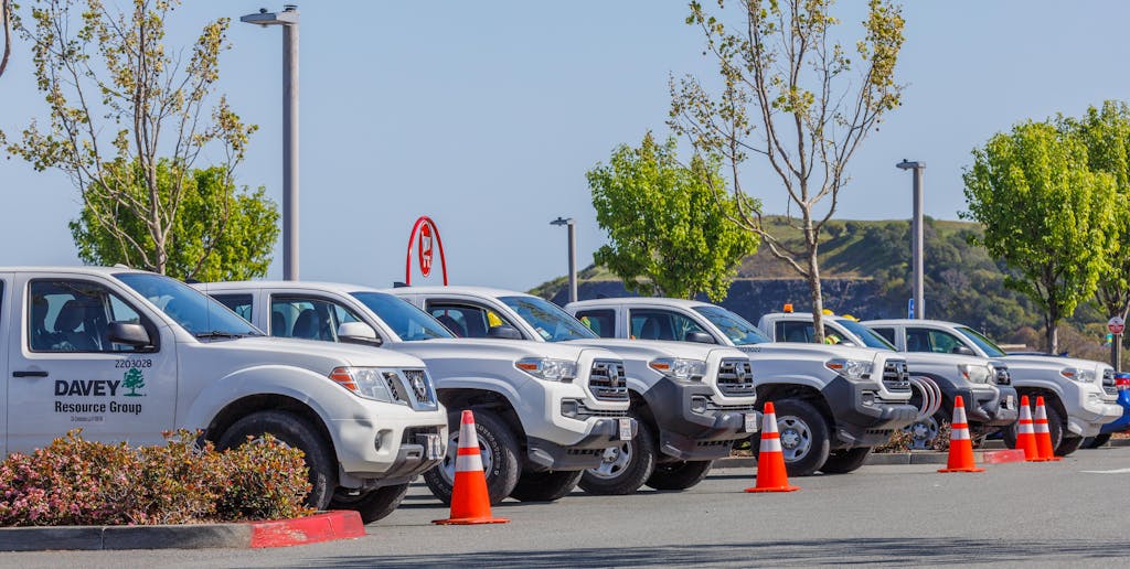 Imagen actual: Fleet of utility trucks parked in an outdoor lot with safety cones and trees.
