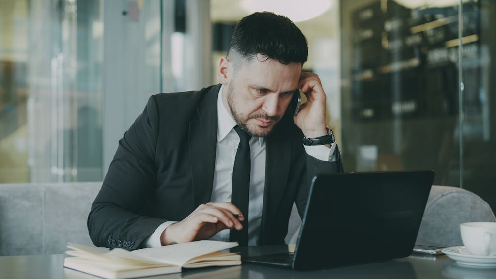 Imagen actual: Focused businessman in suit multitasking on phone and laptop in modern office.