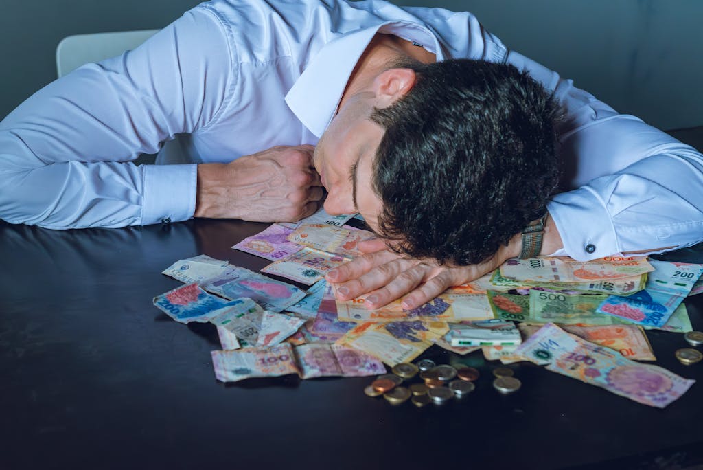 Imagen actual: A tired businessman rests his head on a table covered with Argentine pesos and coins.