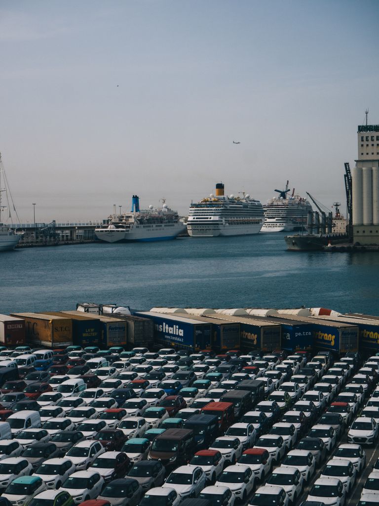 Imagen actual: Cargo and cruise ships docked at Barcelona port with rows of parked cars in foreground.